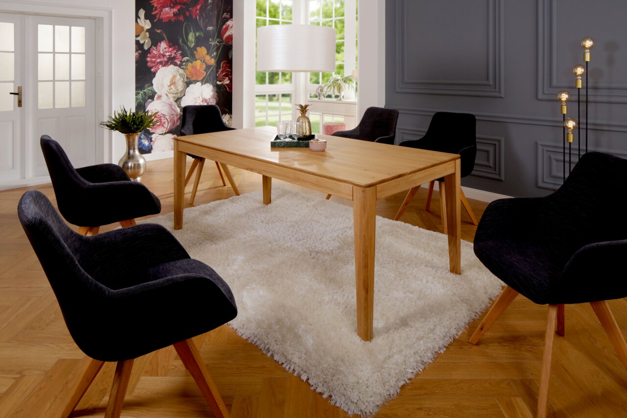 Moderner Esstisch aus Holz in einem stilvollen Esszimmer mit schwarzem Stuhl-Set, Teppich und dekorativer Wand im Hintergrund, seitliche Perspektive.