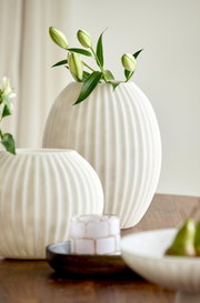 Große weiße, bauchige Vase mit vertikaler Rillenstruktur und grünen Lilienknospen, frontal aus leichter Seitenperspektive auf einem Holztisch arrangiert
