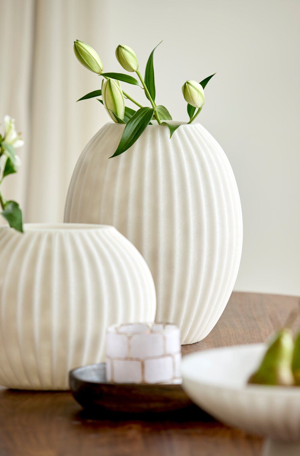Große weiße, bauchige Vase mit vertikaler Rillenstruktur und grünen Lilienknospen, frontal aus leichter Seitenperspektive auf einem Holztisch arrangiert