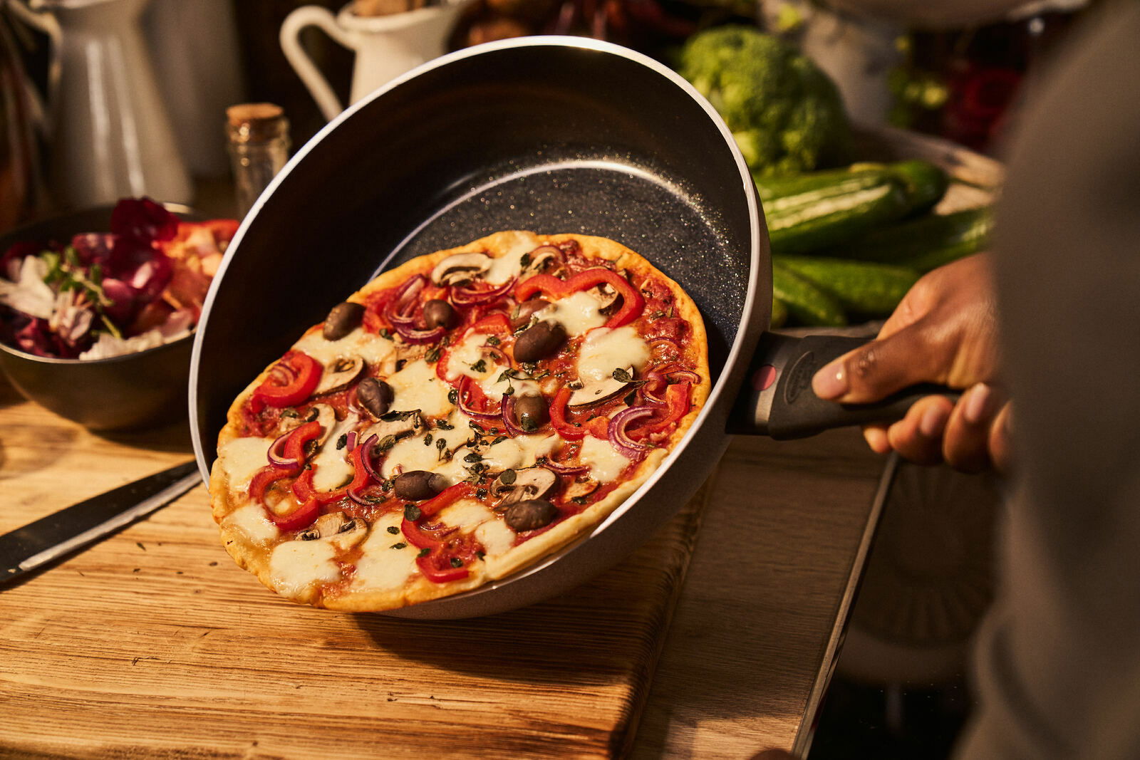 Eine Hand hält eine Bratpfanne mit einer frisch gebackenen Pizza, fotografiert aus einer seitlichen Perspektive.