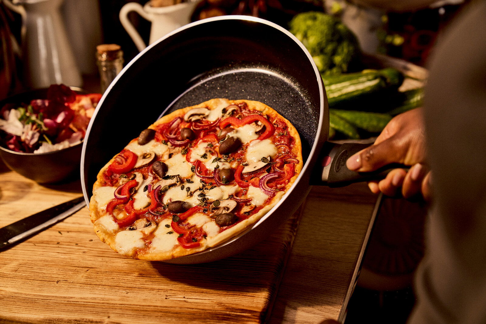 Eine Hand hält eine Bratpfanne mit einer frisch gebackenen Pizza, fotografiert aus einer seitlichen Perspektive.