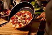 Een hand houdt een koekenpan vast met daarin een versgebakken pizza, gefotografeerd vanuit een zijdelingse hoek.