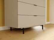 Nahaufnahme einer modernen, cremefarbenen Kommode mit drei Schubladen und schwarzen Griffen, auf einem Holzfußboden stehend.