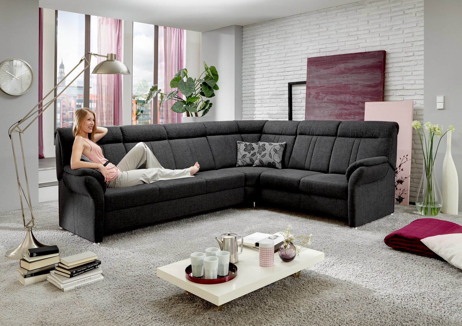 Beldomo Comfort Ecksofa MALAGA Moderne Polsterrundecke in dunklem Grau, perspektivisch von vorne links gezeigt, mit einer entspannten Person darauf sitzend in einem stilvoll eingerichteten Wohnzimmer.