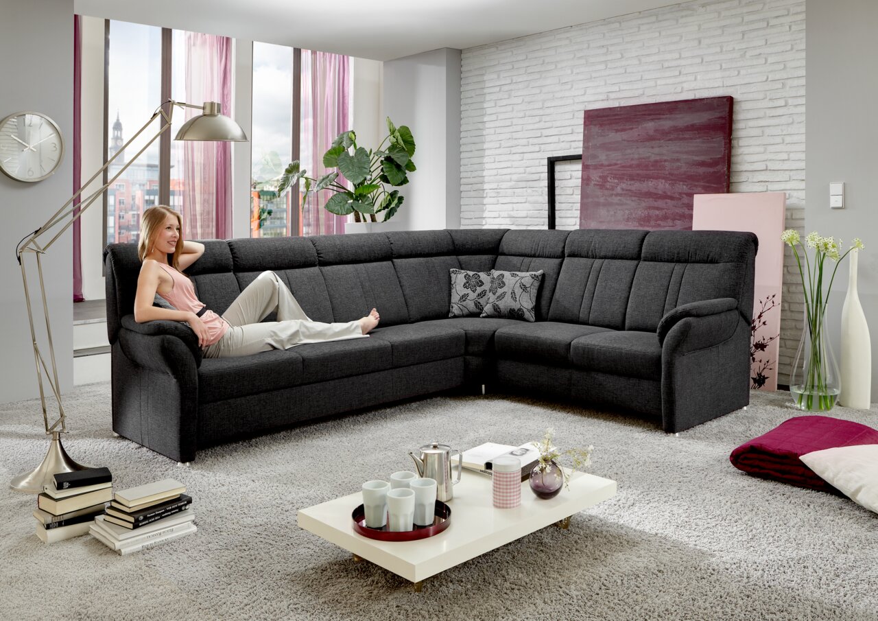 Beldomo Comfort Hoekbank MALAGA Een moderne, gestoffeerde hoekbank in donkergrijs, in perspectief gezien vanaf links vooraan, met daarop een ontspannen persoon in een stijlvol ingerichte woonkamer.