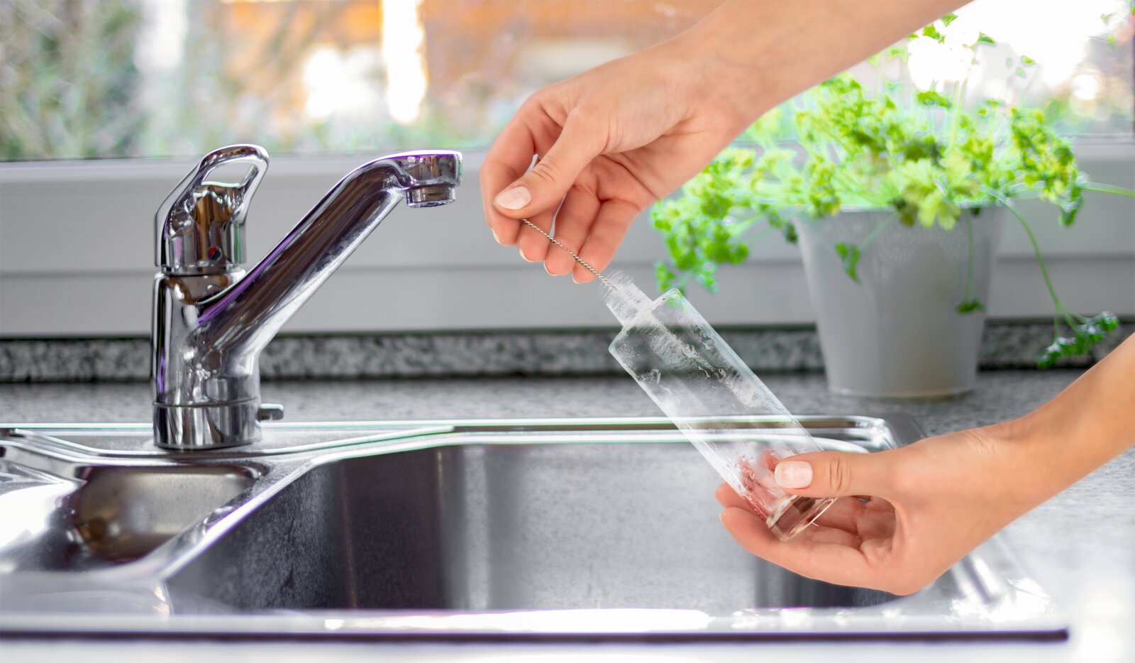 Seitliche Nahaufnahme einer Hand, die eine transparente Trinkflasche mit einer schmalen Reinigungsbürste über einer Küchenspüle säubert