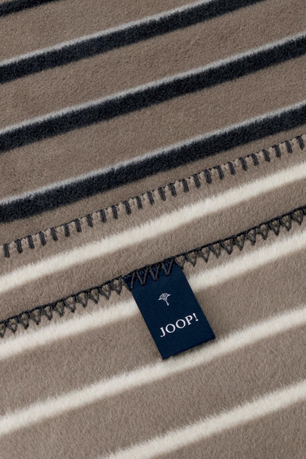 JOOP! Wohndecke 150 x 200 cm Detailaufnahme aus schräger Draufsicht: gestreifte Decke/Plaid in Taupe mit weißen und dunkelgrauen Streifen, Ziernaht am Rand und JOOP!-Label