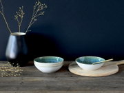 Zwei glasierte Keramikschalen in Weiß mit blau-grünem Innenverlauf auf rustikalem Holztisch, eine auf rundem Holzbrett mit goldenem Löffel, links schwarze Vase mit Zweigen vor dunklem Hintergrund; Perspektive: frontale Seitenansicht auf Tischhöhe.