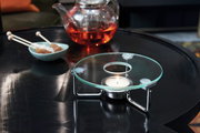 Stövchen mit Teelicht auf einem Tisch, im Hintergrund eine Teekanne mit rotem Tee und eine Schale mit Kandiszucker, seitliche Perspektive.