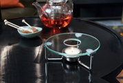 Stövchen mit Teelicht auf einem Tisch, im Hintergrund eine Teekanne mit rotem Tee und eine Schale mit Kandiszucker, seitliche Perspektive.