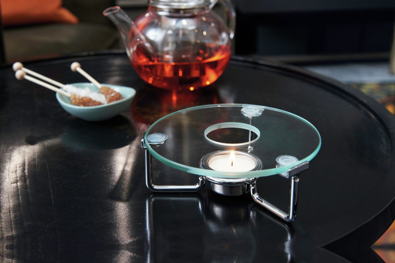 Stövchen mit Teelicht auf einem Tisch, im Hintergrund eine Teekanne mit rotem Tee und eine Schale mit Kandiszucker, seitliche Perspektive.