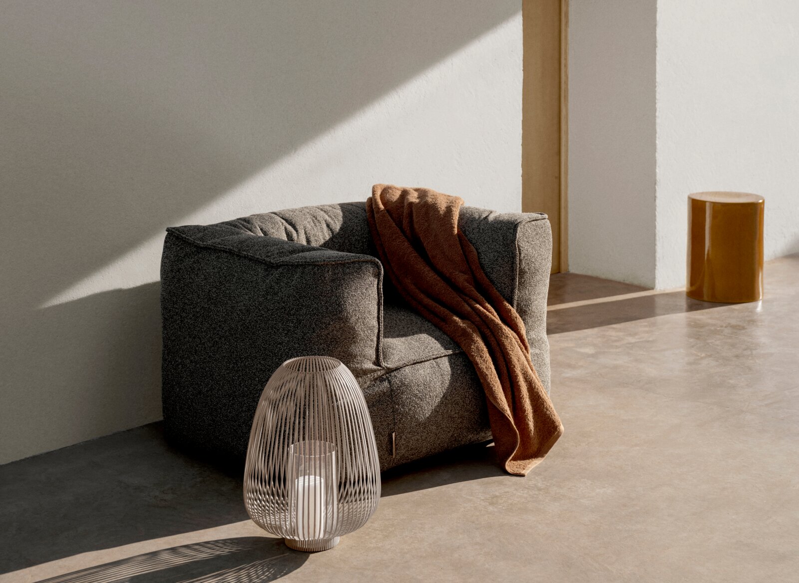 blomus Outdoorfauteuil GROW Moderne grijze loungestoel met brede armleuningen en een bruine deken, met daarvoor een witte draadlantaarn met kaars; foto genomen vanuit een licht schuin vooraanzicht in een minimalistische ruimte met warm zonlicht.