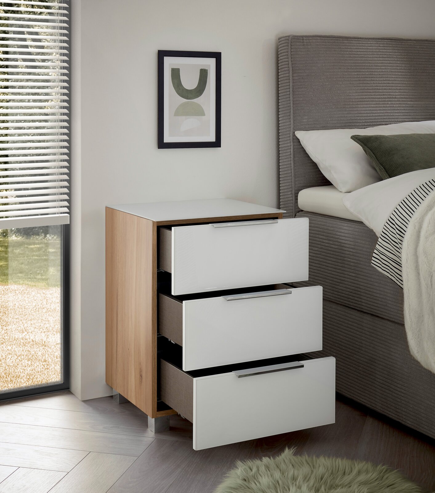Moderne Nachtkommode mit drei offenen Schubladen, Holz- und Weißlack-Design, seitliche Perspektive neben einem Bett.