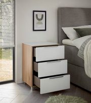 Moderne Nachtkommode mit drei offenen Schubladen, Holz- und Weißlack-Design, seitliche Perspektive neben einem Bett.