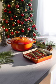 Rote Sauteuse und Auflaufform mit Tomaten und Rosmarin auf einem Tisch, im Hintergrund ein geschmückter Weihnachtsbaum. Perspektive von schräg oben.