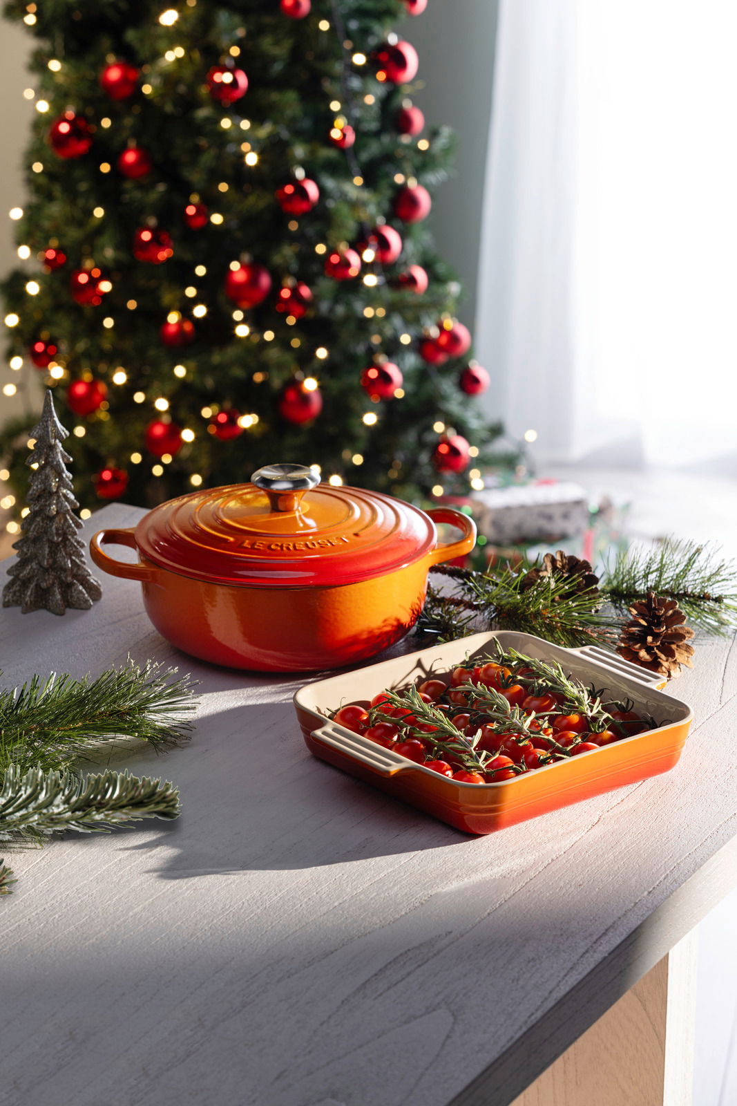 Rote Sauteuse und Auflaufform mit Tomaten und Rosmarin auf einem Tisch, im Hintergrund ein geschmückter Weihnachtsbaum. Perspektive von schräg oben.