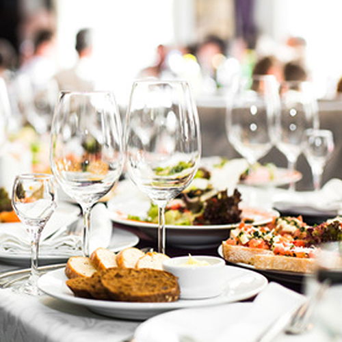 Gedeckter Tisch mit Brot, Gläsern und Speisen in einem belebten Raum.