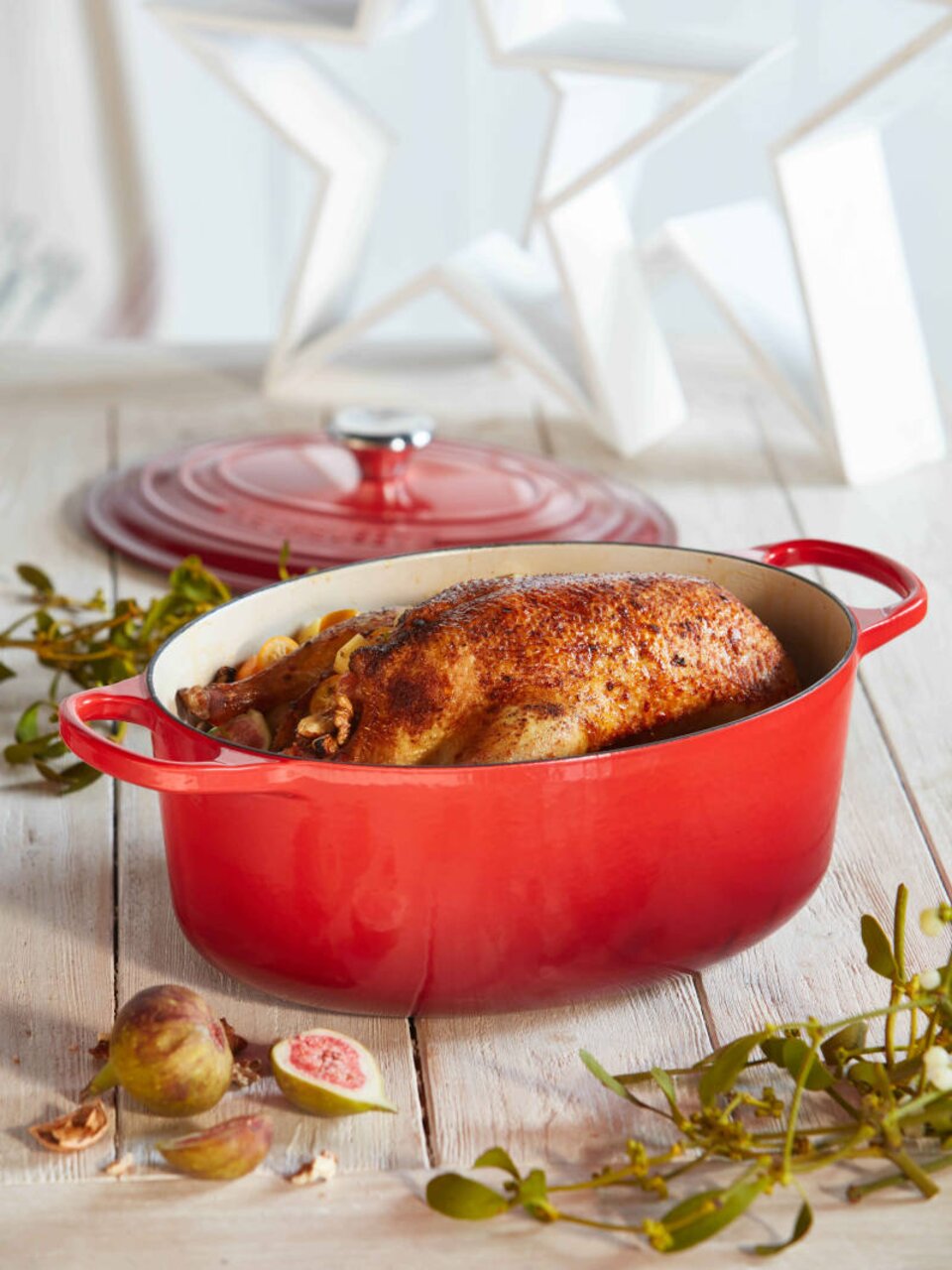 Roter ovaler Bräter mit Deckel, gefüllt mit einem gebratenen Hähnchen, auf einem Holztisch mit Feigen und Zweigen, seitliche Perspektive.