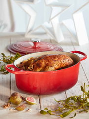 Roter ovaler Bräter mit Deckel, gefüllt mit einem gebratenen Hähnchen, auf einem Holztisch mit Feigen und Zweigen, seitliche Perspektive.