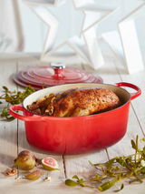 Roter ovaler Bräter mit Deckel, gefüllt mit einem gebratenen Hähnchen, auf einem Holztisch mit Feigen und Zweigen, seitliche Perspektive.