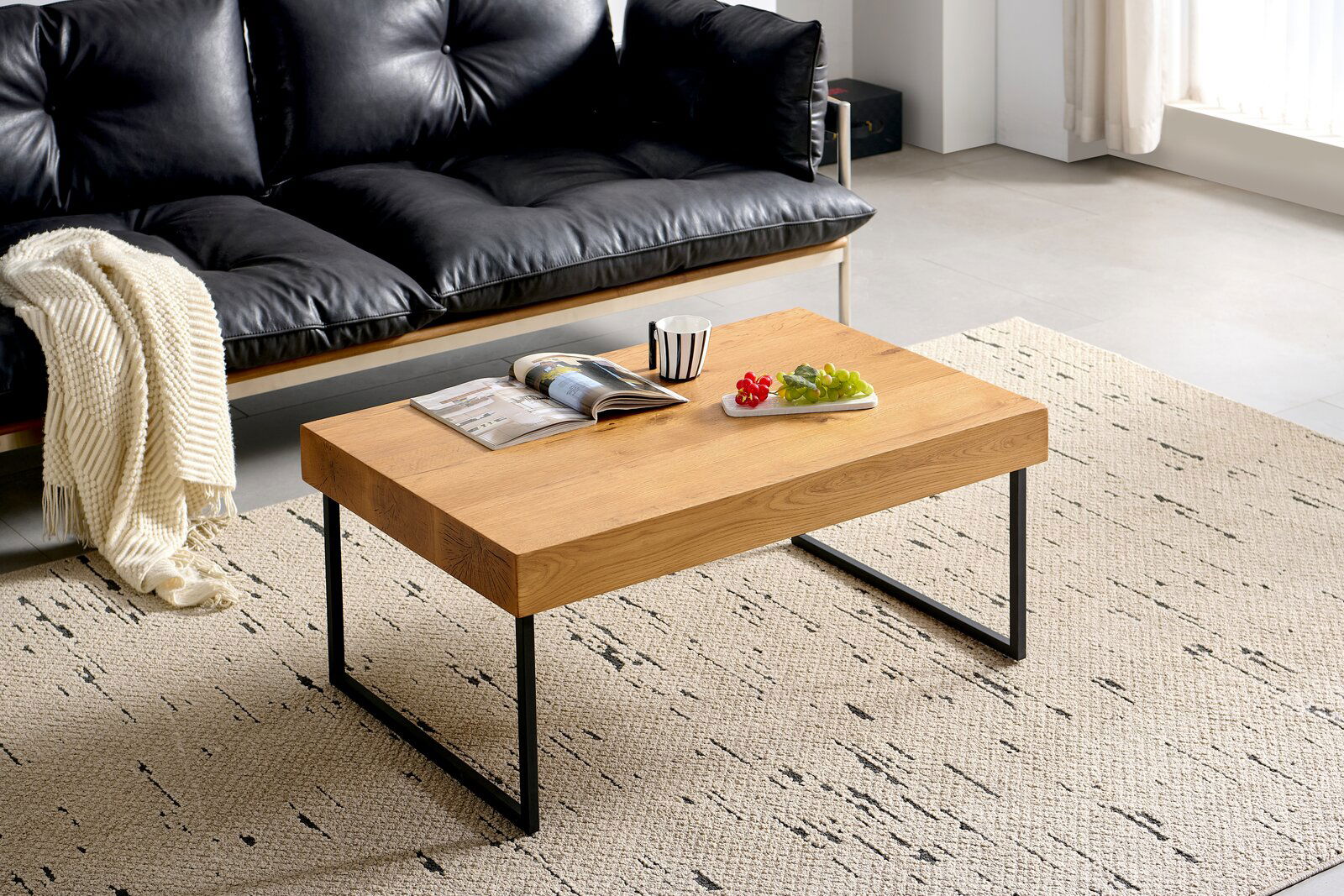 Moderner Couchtisch aus Holz mit Metallbeinen, von schräg oben fotografiert, auf einem gemusterten Teppich vor einem schwarzen Ledersofa.