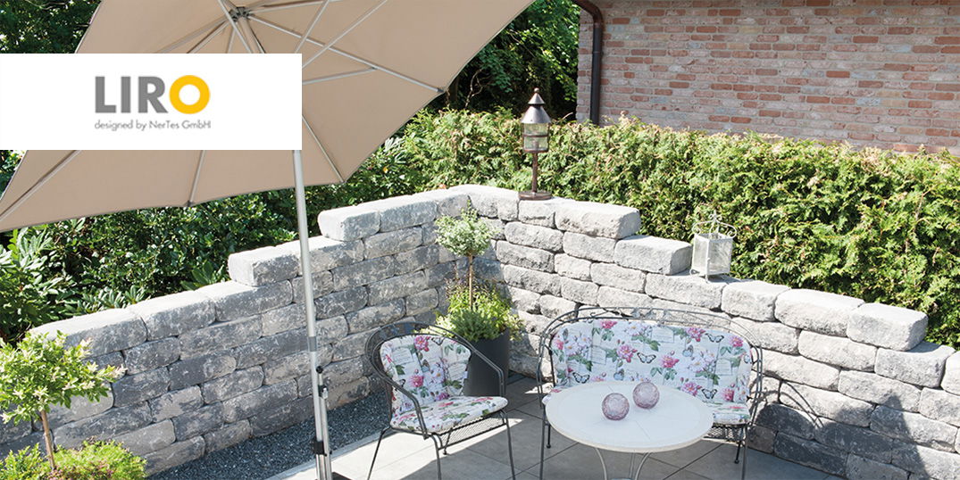 Kleiner Sitzbereich im Freien vor einer rustikalen Steinwand und einem großen Sonnenschirm der Marke Liro.