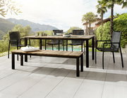 Moderner Gartentisch und Gartenbank aus Holz und Metall auf einer Terrasse mit Blick auf die Berge, umgeben von Palmen und grüner Landschaft. Perspektive von schräg vorne.
