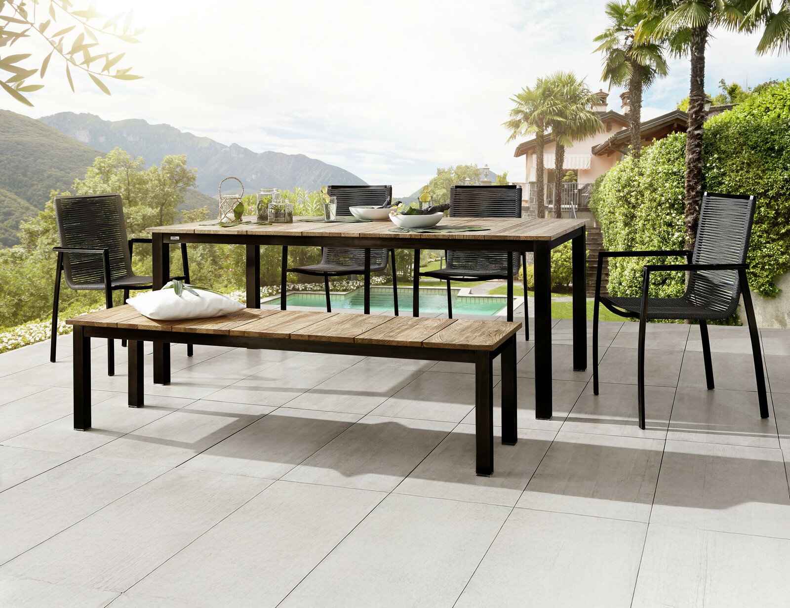 Amatio Tuintafel JAEN Moderner Gartentisch und Gartenbank aus Holz und Metall auf einer Terrasse mit Blick auf die Berge, umgeben von Palmen und grüner Landschaft. Perspektive von schräg vorne.