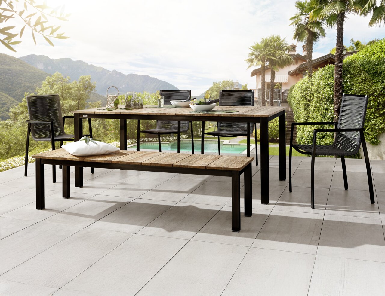 Amatio Gartenbank JAEN Moderner Gartentisch und Gartenbank aus Holz und Metall auf einer Terrasse mit Blick auf die Berge, umgeben von Palmen und grüner Landschaft. Perspektive von schräg vorne.