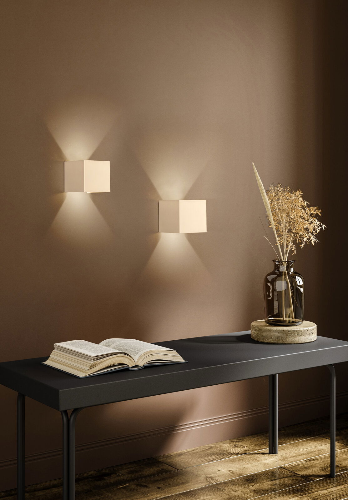 Moderne weiße kubische Wandleuchten mit Up- und Downlight an brauner Wand über schwarzer Konsole mit offenem Buch und Glasvase mit Trockenblumen, Perspektive: frontale Ansicht auf Augenhöhe