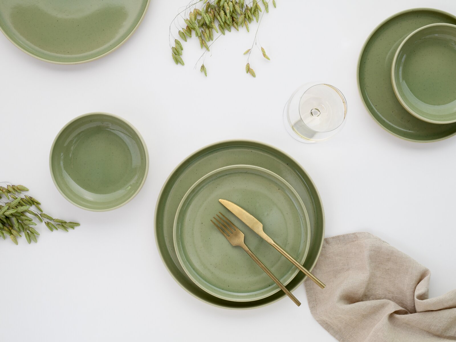 Draufsicht (Flatlay) auf ein gedecktes Arrangement mit grün gesprenkeltem Keramikgeschirr (Teller und Schalen), goldenem Besteck, Weinglas, Leinenserviette und dekorativen Zweigen auf weißem Hintergrund.
