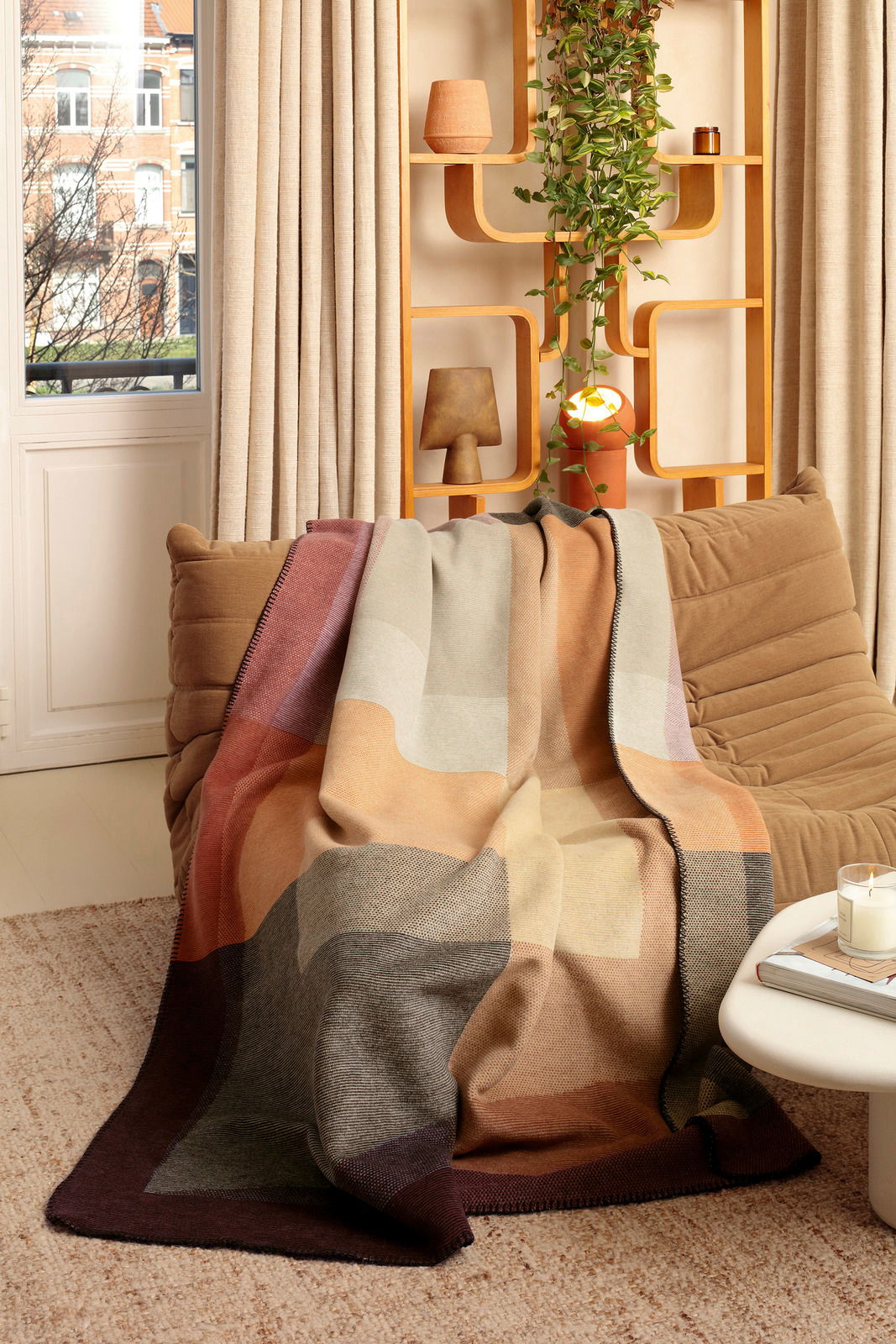 Gemütliche Wohndecke in warmen Erdtönen auf einem beigen Sofa, mit dekorativem Regal im Hintergrund. Perspektive: Frontalansicht.