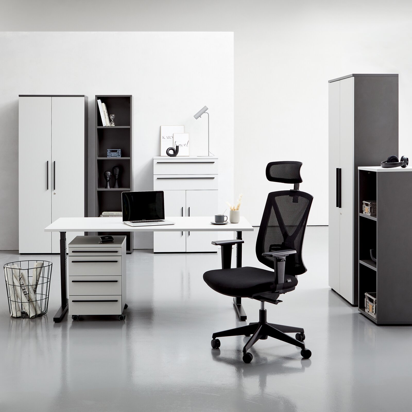 Moderne Büromöbel in einem hellen Raum, darunter ein Winkelschreibtisch, Rollcontainer, Aktenschränke und ein ergonomischer Bürostuhl. Frontalansicht.