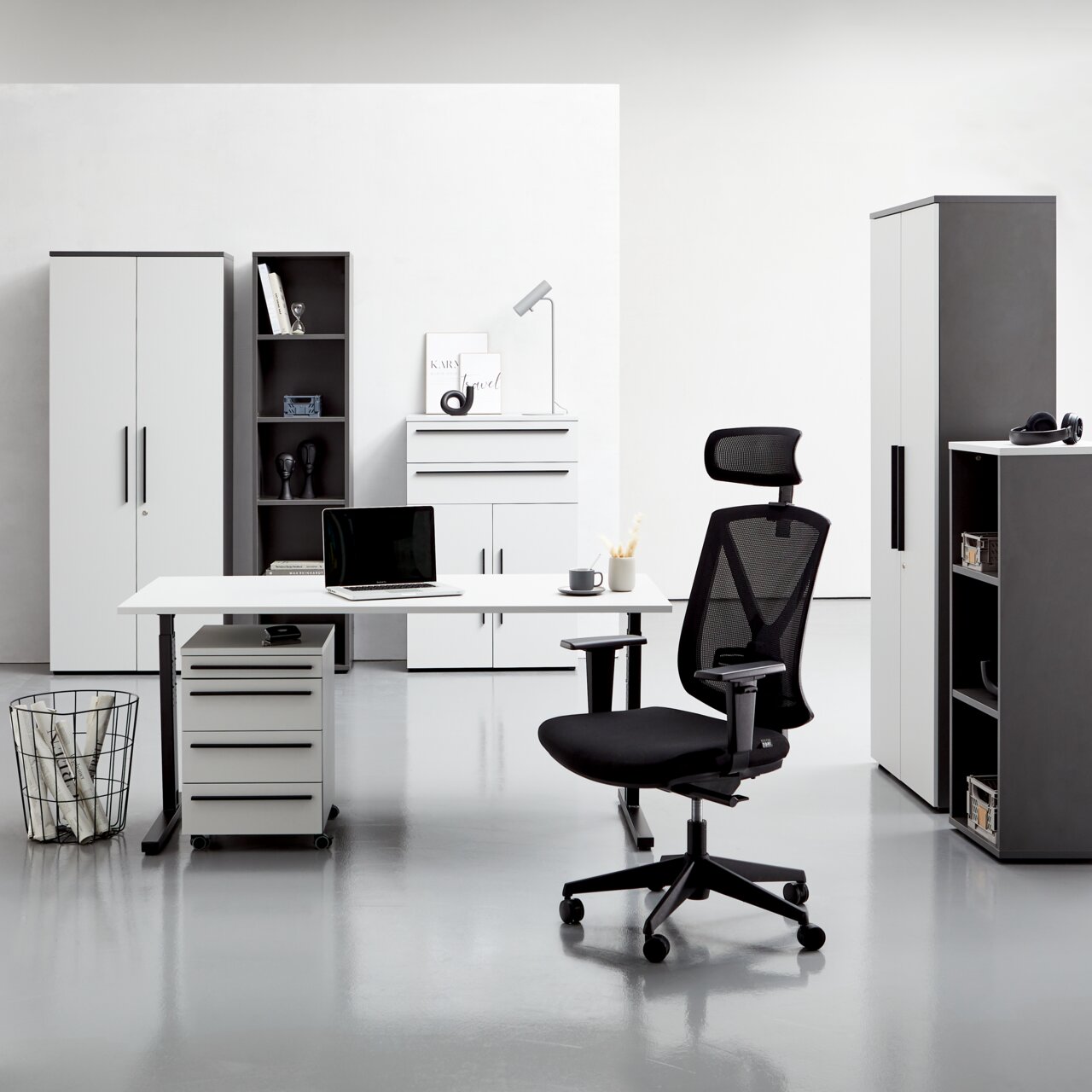 Moderne Büromöbel in einem hellen Raum, darunter ein Winkelschreibtisch, Rollcontainer, Aktenschränke und ein ergonomischer Bürostuhl. Frontalansicht.