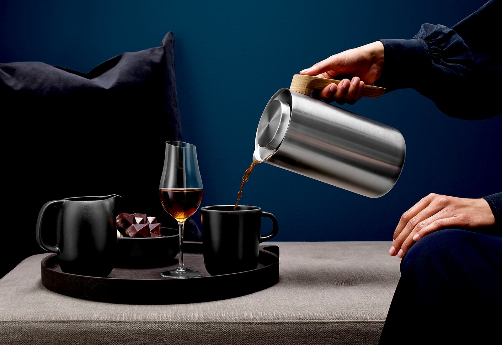 Schuin zijaanzicht van een roestvrijstalen thermoskan met houten handgreep die koffie inschenkt in een zwarte mok op een dienblad, met een glas en karaf op een bank.