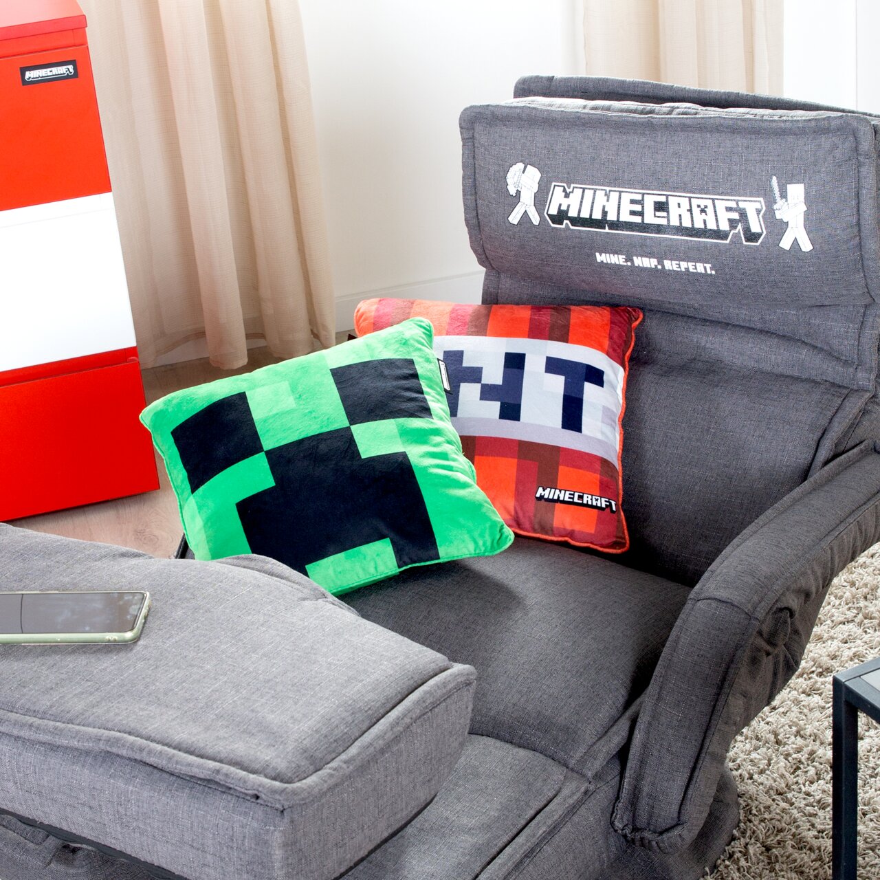 Grijze Minecraft-gamestoel met geborduurde rugleuning „MINECRAFT – Mine. Naa. Repeat.“ en twee Minecraft-kussens (Creeper en pixelmotief), gefotografeerd in een schuin vooraanzicht van bovenaf.