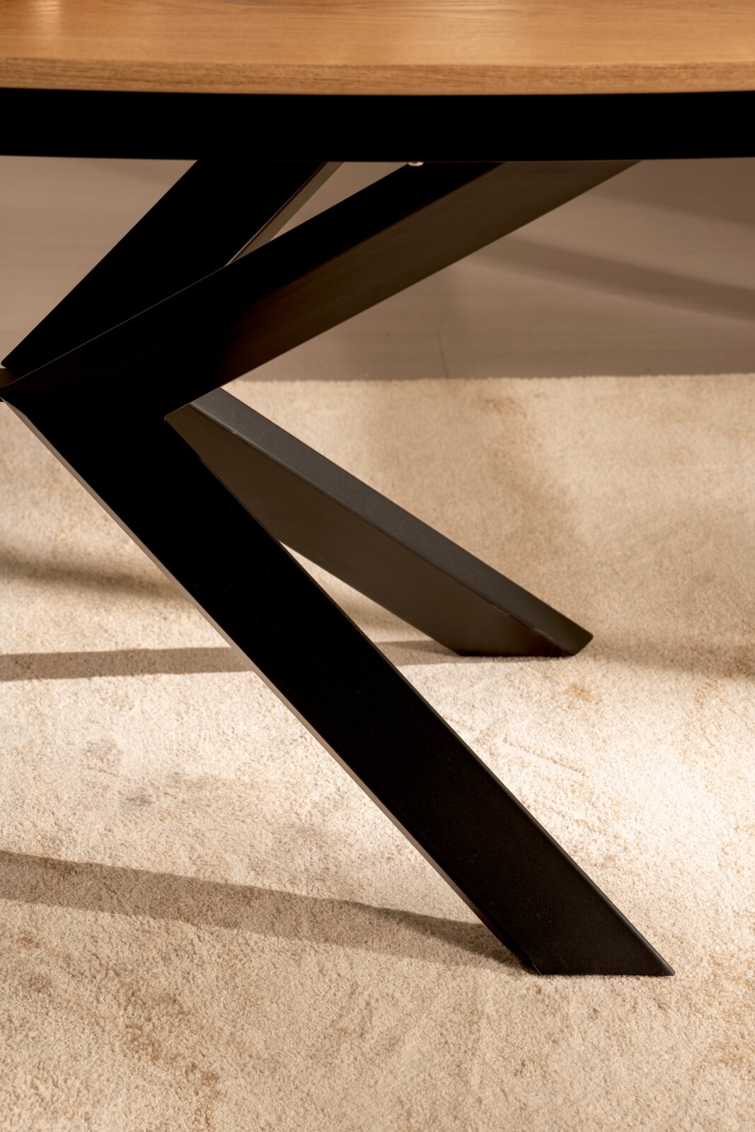 Detailaufnahme in leichter Untersicht: schwarzes, X-förmiges Metall-Tischgestell unter einer Holz-Tischplatte auf hellem Teppich