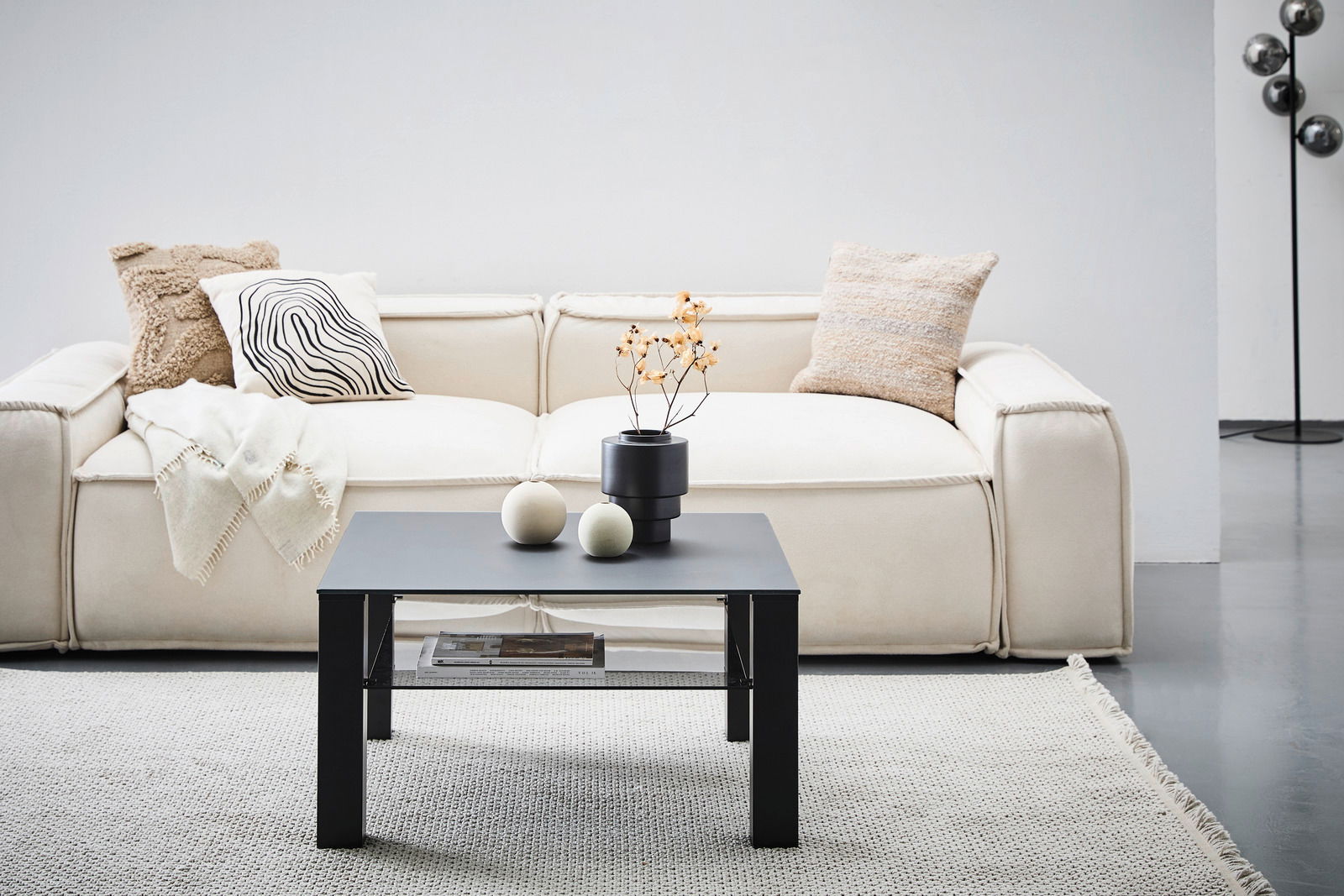 Moderner schwarzer Beistelltisch aus Metall, frontal vor einem cremefarbenen Sofa mit dekorativen Kissen und einer Decke, auf einem hellen Teppich.