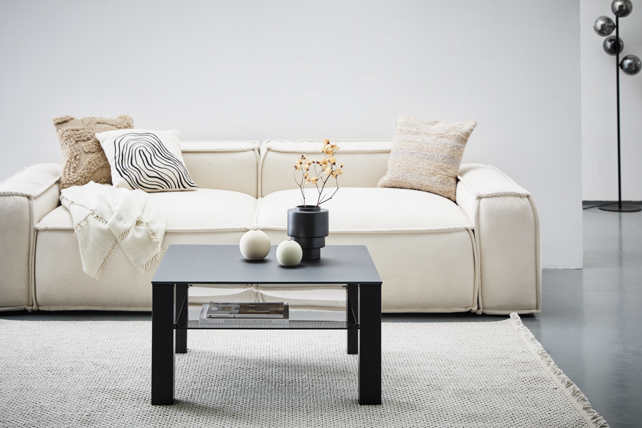 Moderner schwarzer Beistelltisch aus Metall, frontal vor einem cremefarbenen Sofa mit dekorativen Kissen und einer Decke, auf einem hellen Teppich.