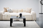 Moderner schwarzer Beistelltisch aus Metall, frontal vor einem cremefarbenen Sofa mit dekorativen Kissen und einer Decke, auf einem hellen Teppich.