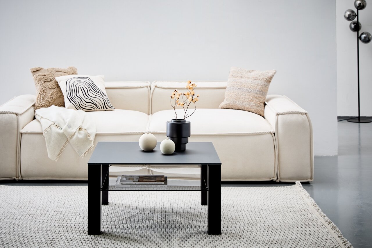 Moderner schwarzer Beistelltisch aus Metall, frontal vor einem cremefarbenen Sofa mit dekorativen Kissen und einer Decke, auf einem hellen Teppich.