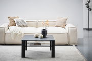 Moderner schwarzer Beistelltisch aus Metall, frontal vor einem cremefarbenen Sofa mit dekorativen Kissen und einer Decke, auf einem hellen Teppich.