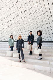 Vier Frauen stehen auf einer Treppe vor einer modernen, geometrischen Wand und halten jeweils eine cremefarbene Tasche in der Hand. Die Perspektive ist frontal.