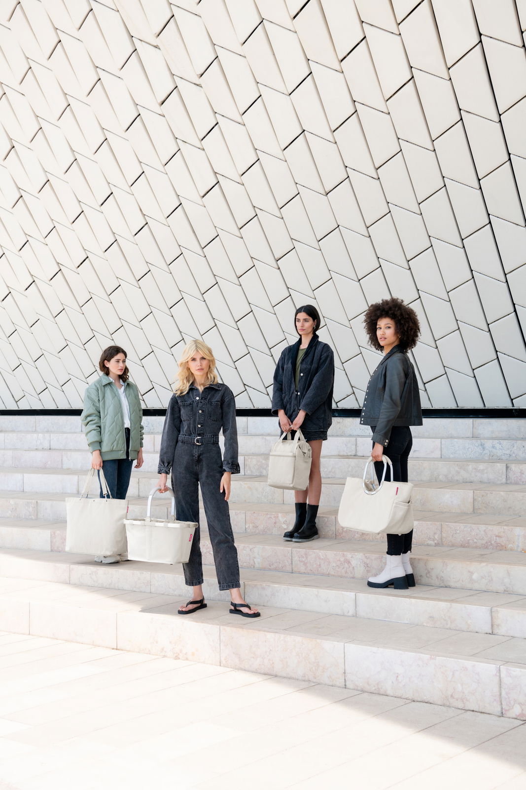 Vier Frauen stehen auf einer Treppe vor einer modernen, geometrischen Wand und halten jeweils eine cremefarbene Tasche in der Hand. Die Perspektive ist frontal.