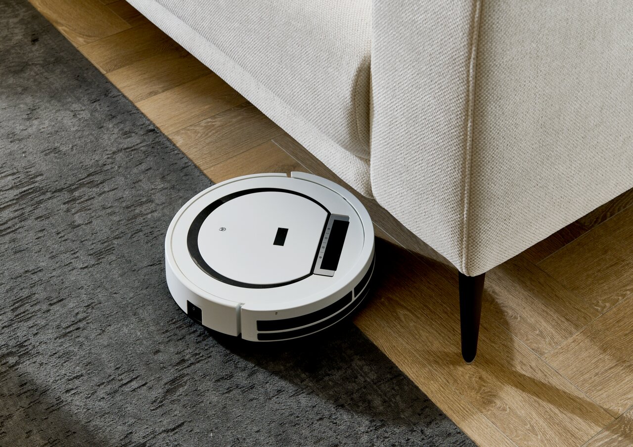 Een witte robotstofzuiger maakt de vloer schoon naast een lichte hoekbank, gezien vanuit vogelperspectief.