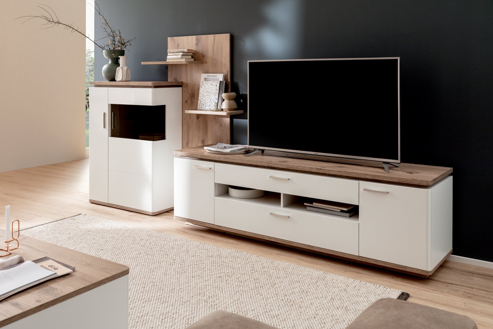 Modernes Wohnzimmer-Set in Weiß mit Holzoptik: TV-Lowboard mit Schubladen und offenen Fächern sowie passendes Highboard und Wandregal, schräg von vorne aus leicht seitlicher Perspektive fotografiert.