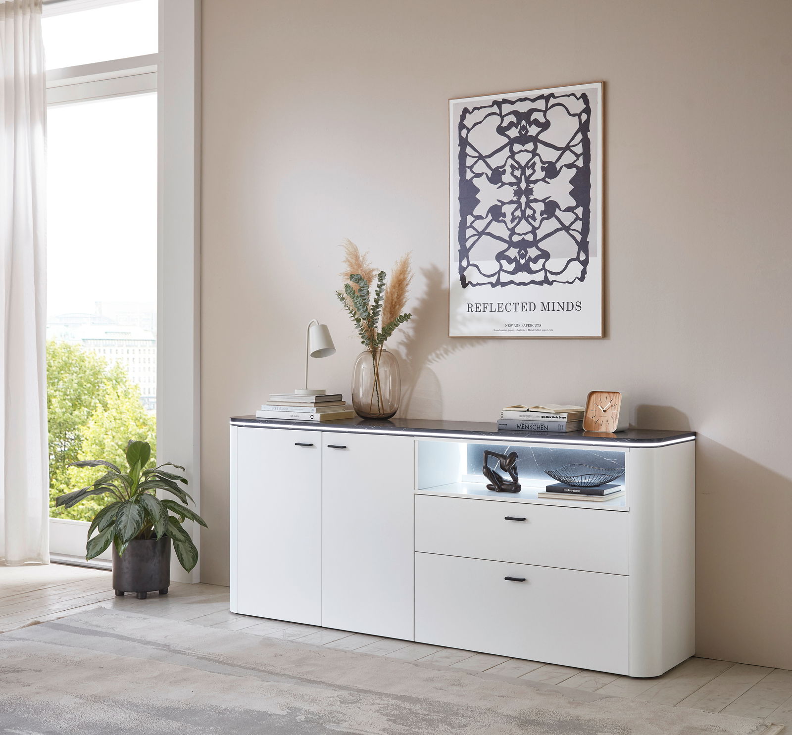 Zurbrüggen Dressoir WAVE Modernes weißes Sideboard mit zwei Türen und zwei Schubladen, dekoriert mit einer Vase, Büchern und einer Lampe, in einem hellen Raum mit großem Fenster, seitliche Perspektive.