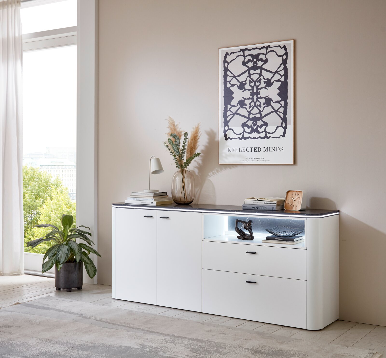 Modernes weißes Sideboard mit zwei Türen und zwei Schubladen, dekoriert mit einer Vase, Büchern und einer Lampe, in einem hellen Raum mit großem Fenster, seitliche Perspektive.