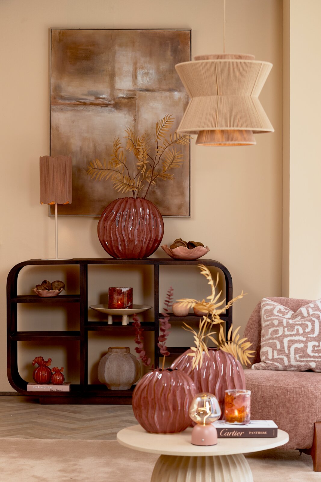 Frontaal aanzicht van een sfeervol interieur in warme aardetinten met een donkerhouten vakkenkast, roze glazen vazen, zachte fauteuil en elegante koordlampen.