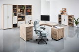 Moderne Büroumgebung mit Eckschreibtisch, Rollcontainer und Regalen in Holzoptik, aus der Perspektive eines Betrachters im Raum.